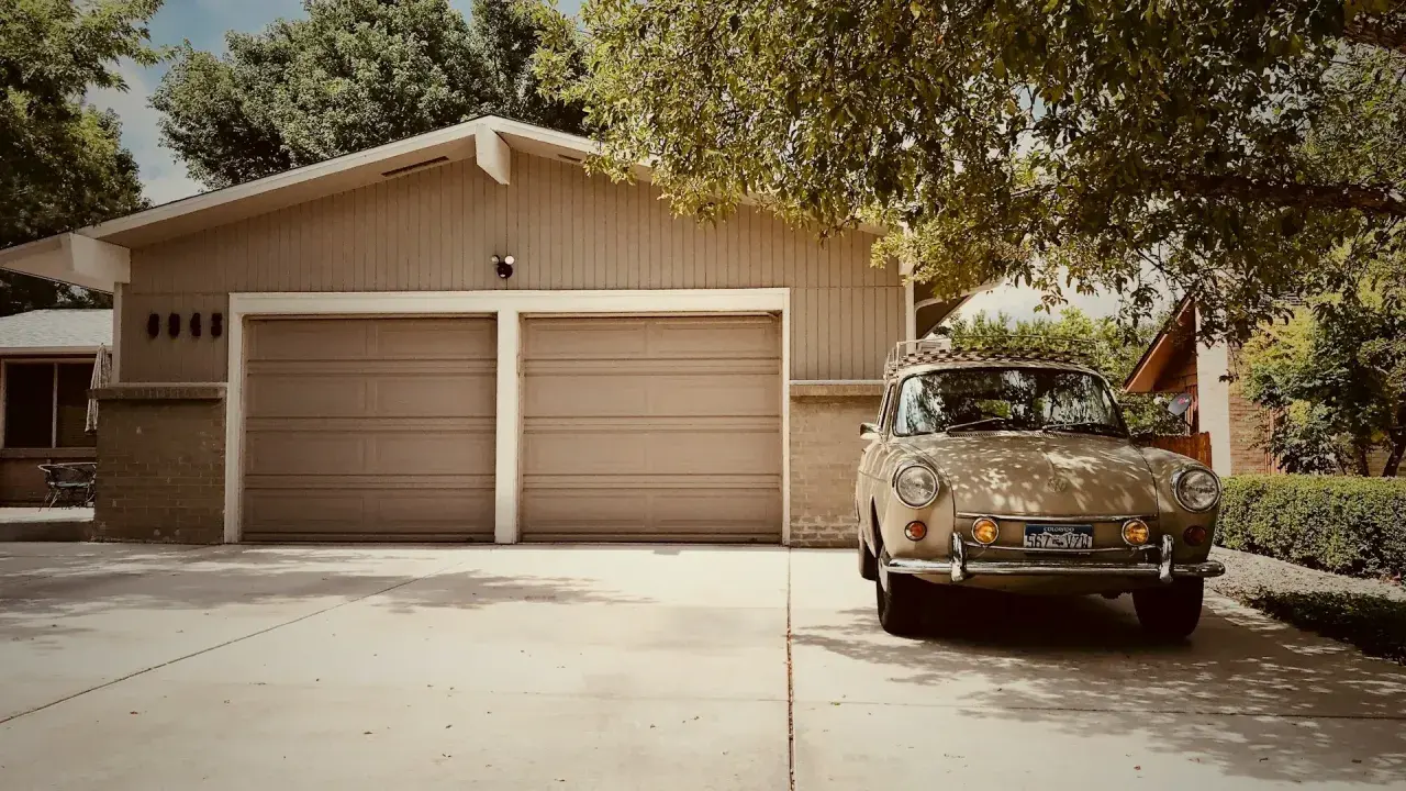 En beige veteranbil står parkerad framför ett garage med två portar. Kanske är det dags att bygga garage för att skydda den?