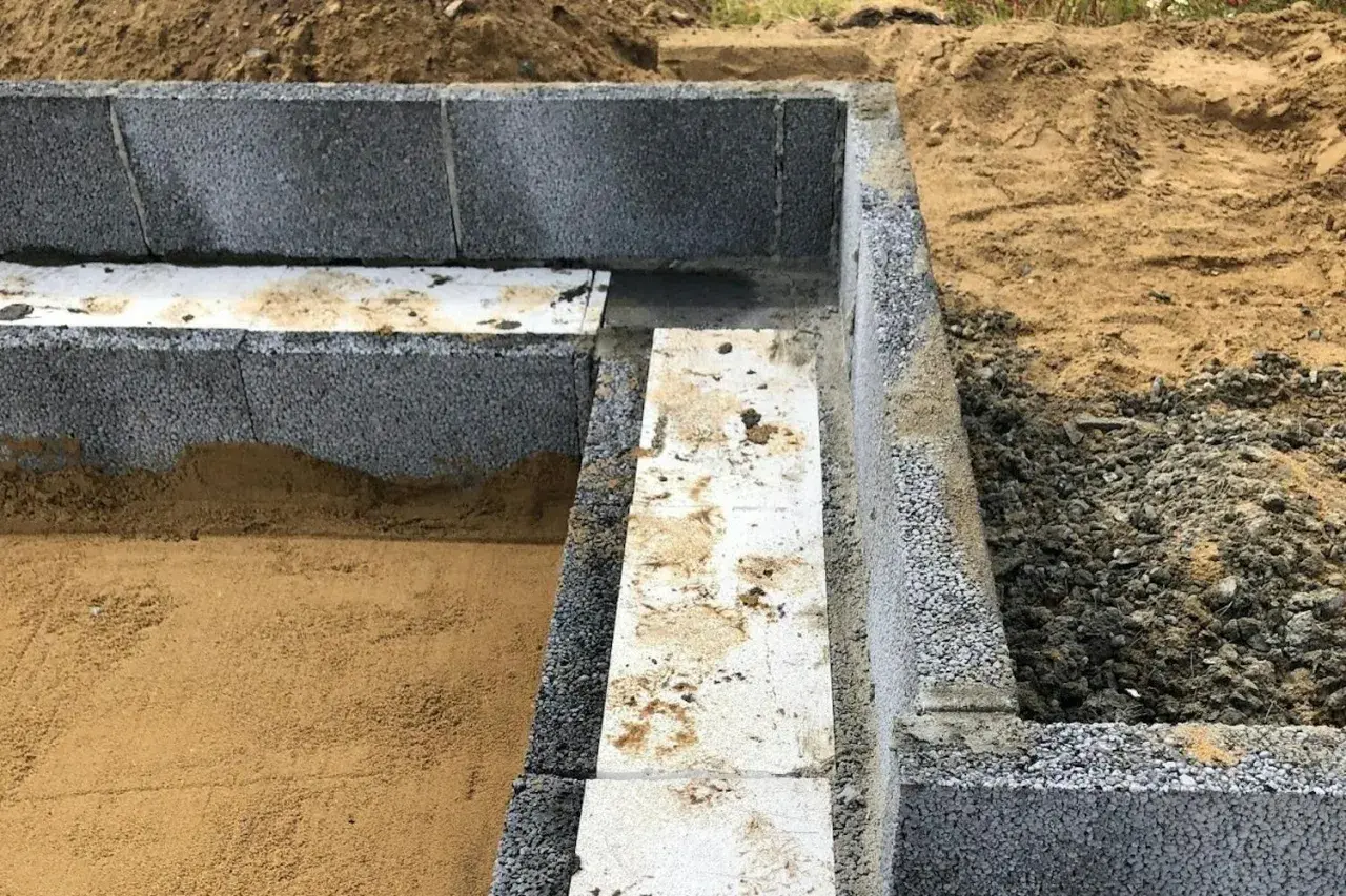 Grundläggande arbete med lecablock. En del av grunden är färdigbyggd med grå lecablock och isolering. Sand och jord syns runt omkring.