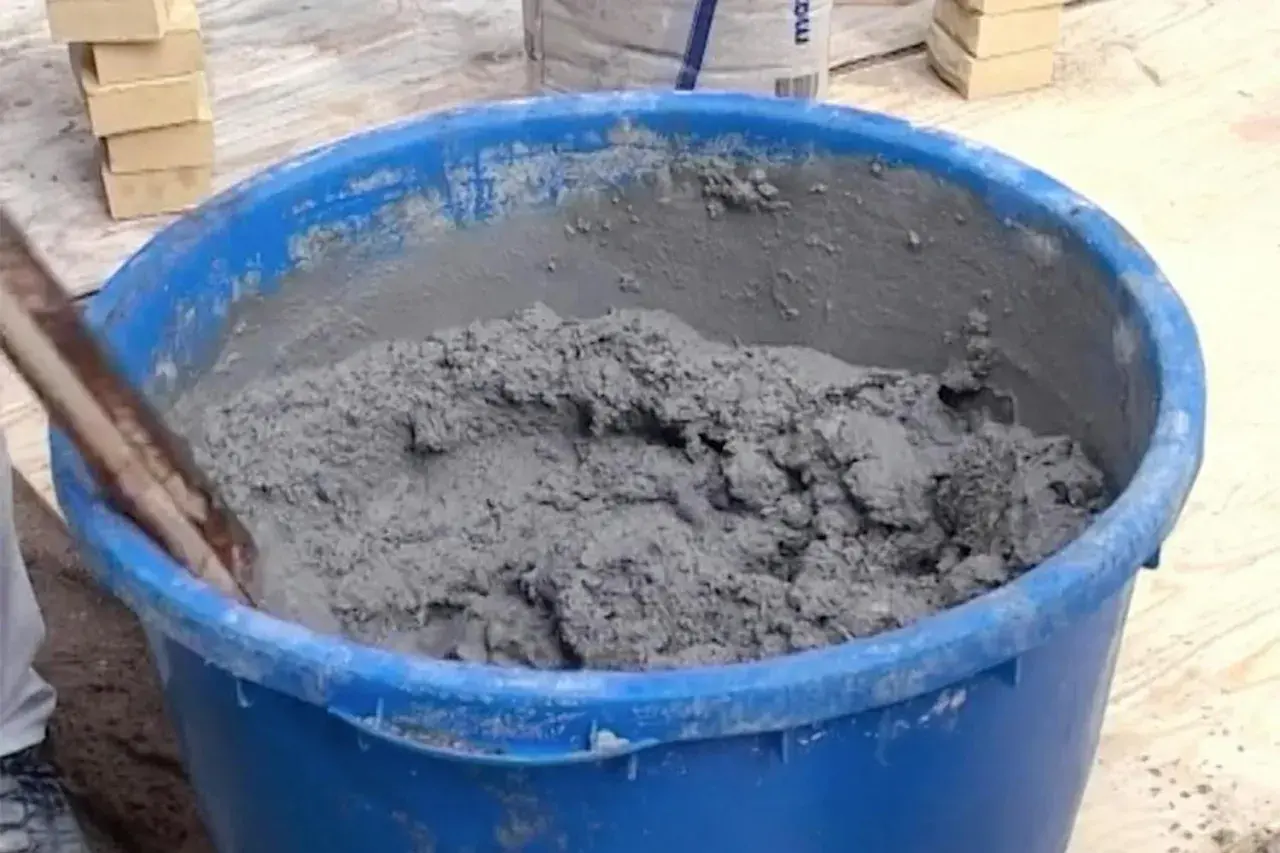 En blå balja fylld med grå cement som någon håller på att blanda. Tegelstenar syns i bakgrunden.