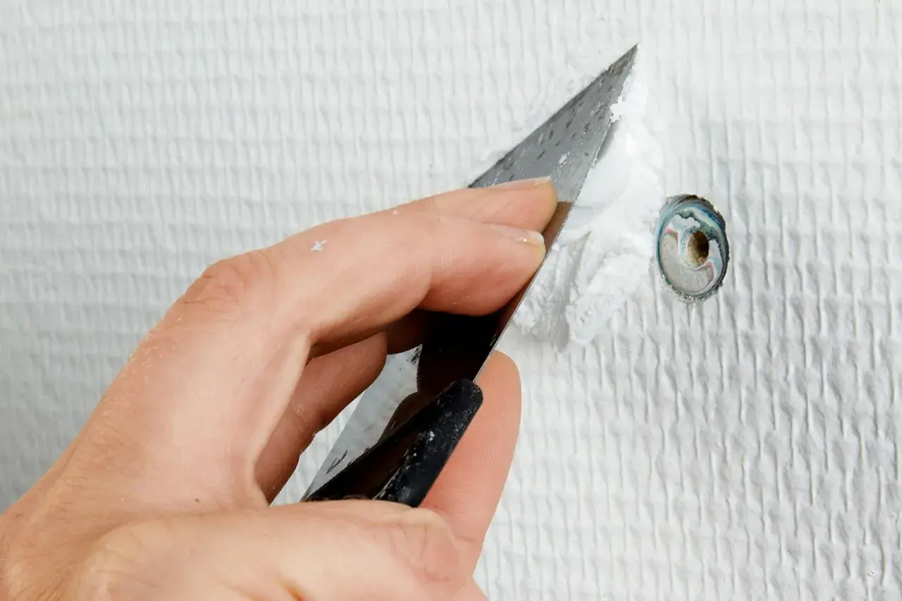 Hand spacklar ett hål i en vit, strukturerad vägg med en spackel.
