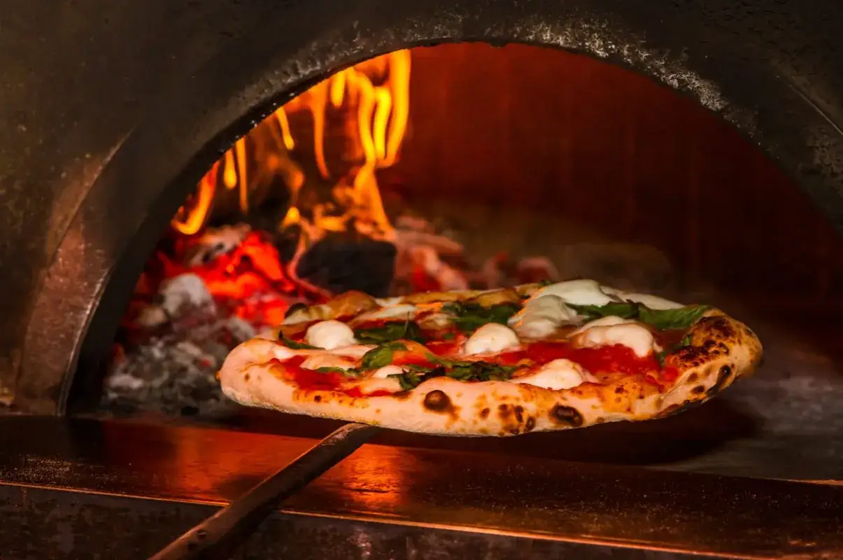 En pizza med mozzarella och basilika tas ur en vedeldad pizzaugn. Flammor syns i bakgrunden.