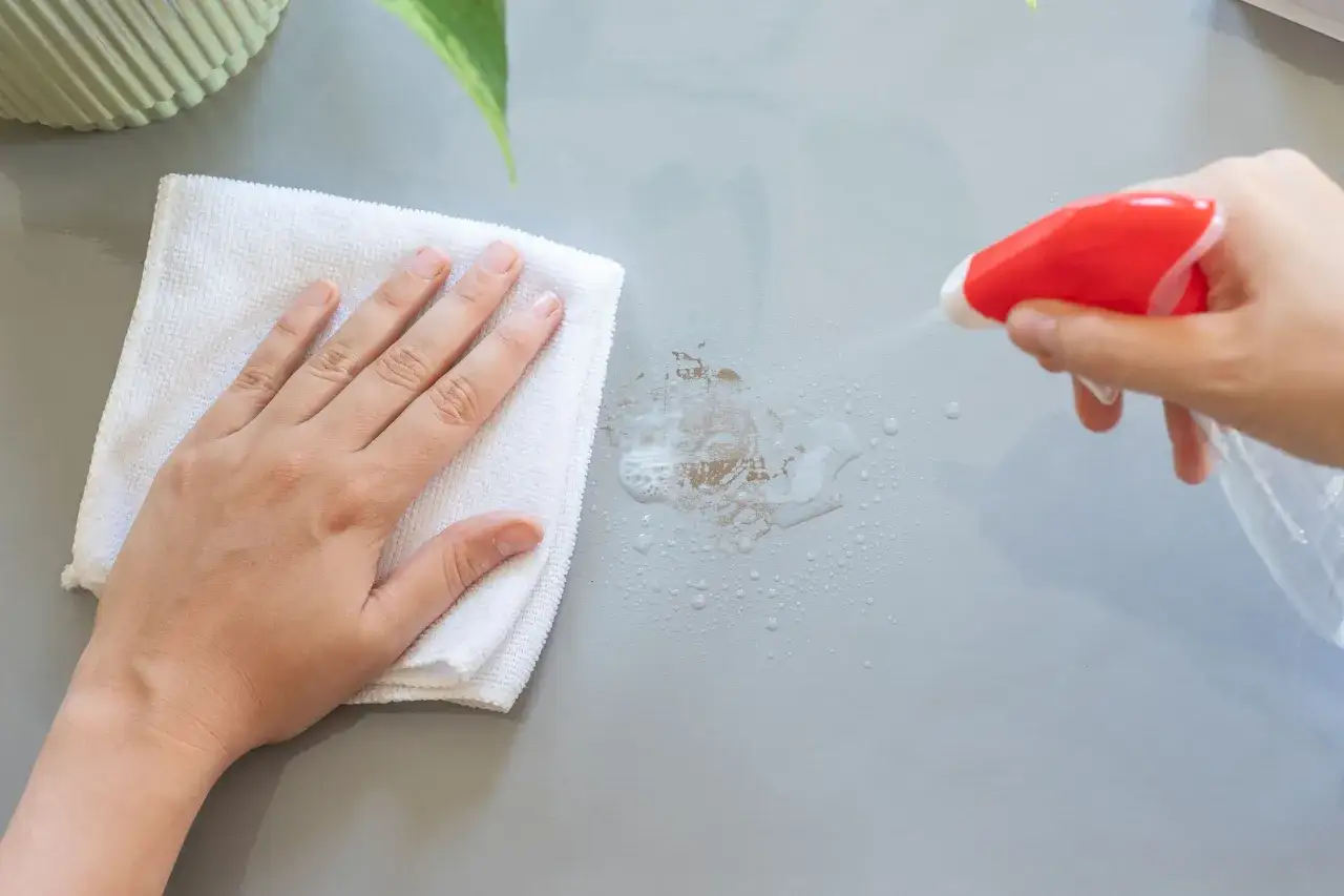 En hand med en vit trasa torkar bort envisa tejprester fr&aring;n en gr&aring; yta. En sprayflaska sprutar reng&ouml;ringsv&auml;tska.