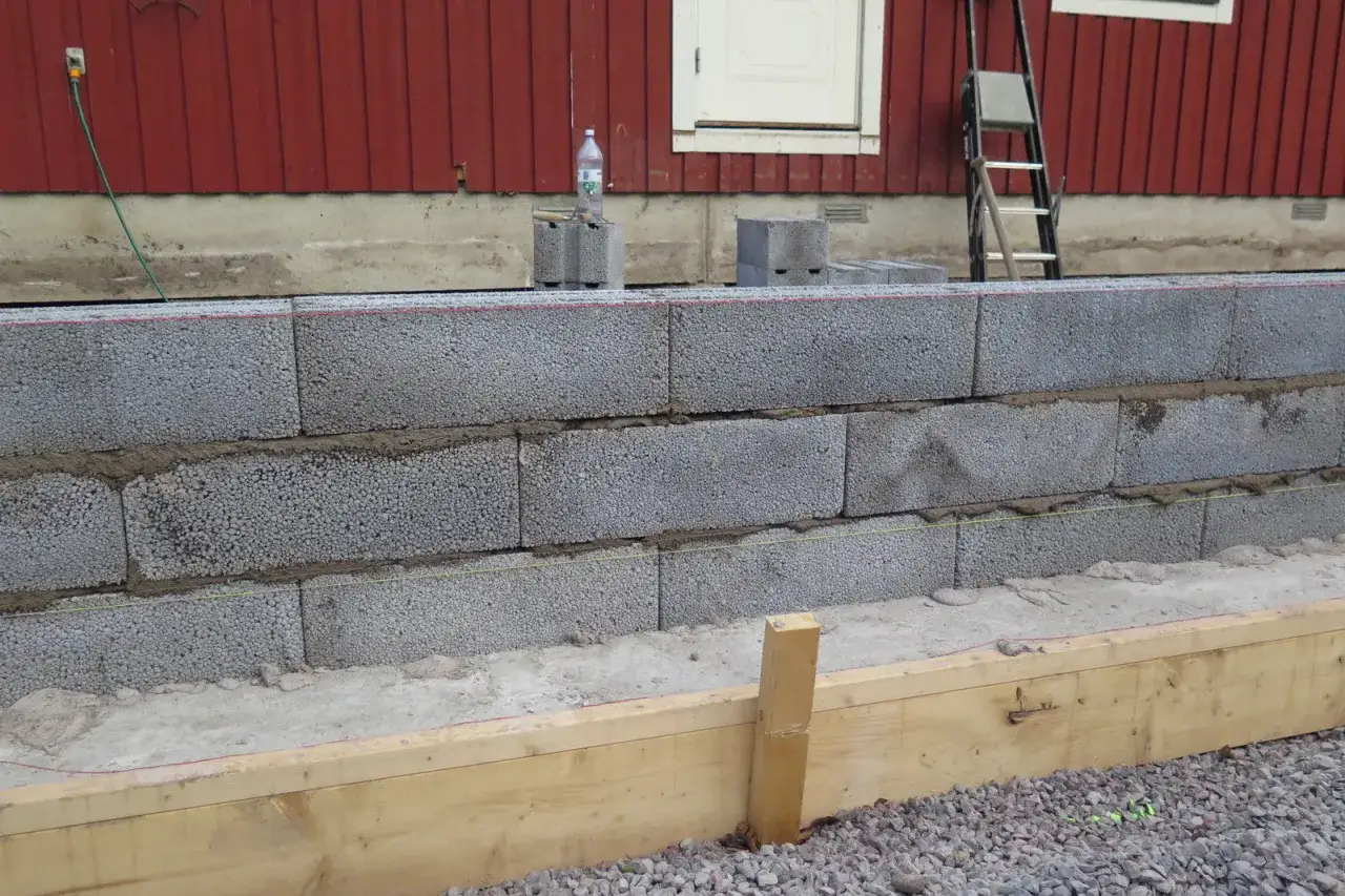 Byggnation av en mur av lecablock framf&ouml;r ett r&ouml;tt tr&auml;hus. Muren &auml;r i flera lager med cement emellan.