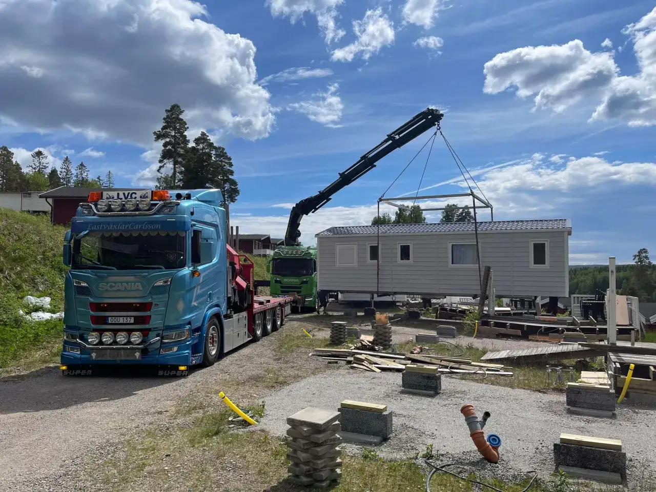 En kranlyft f&ouml;r att flytta friggebod. En bl&aring; Scania-lastbil med kran och en gr&ouml;n lastbil med huset.