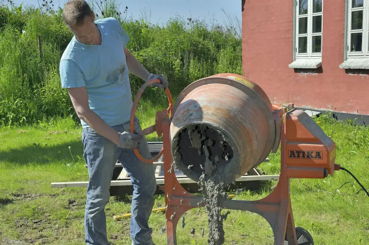 Man i bl&aring; tr&ouml;ja och jeans anv&auml;nder en cementblandare f&ouml;r att blanda cement utomhus.