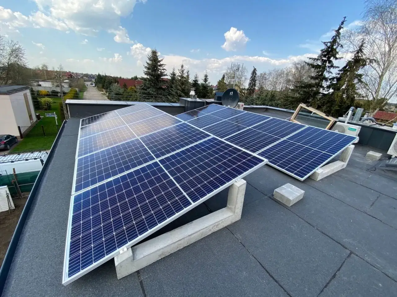 Solceller p&aring; ett platt tak f&aring;ngar solens energi. En gr&ouml;nskande omgivning och bl&aring; himmel med moln.