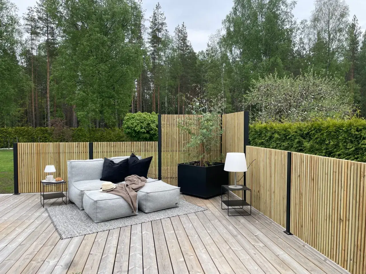 En mysig uteplats med gr&aring; soffa, svarta kuddar och en filt. En st&aring;ende plank ribb-staket ramar in.