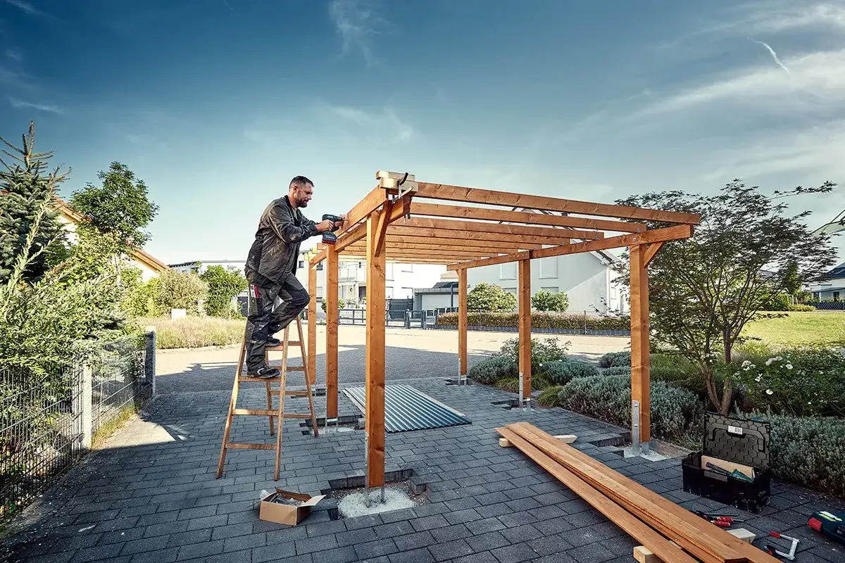 Man bygger carport med tr&auml;stomme och skruvdragare. Solig dag med bl&aring; himmel.