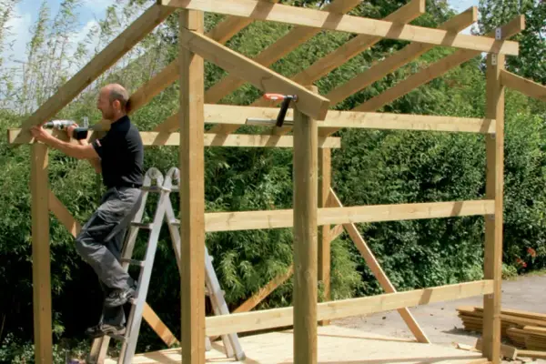 Man on ladder building wooden shed frame with drill.